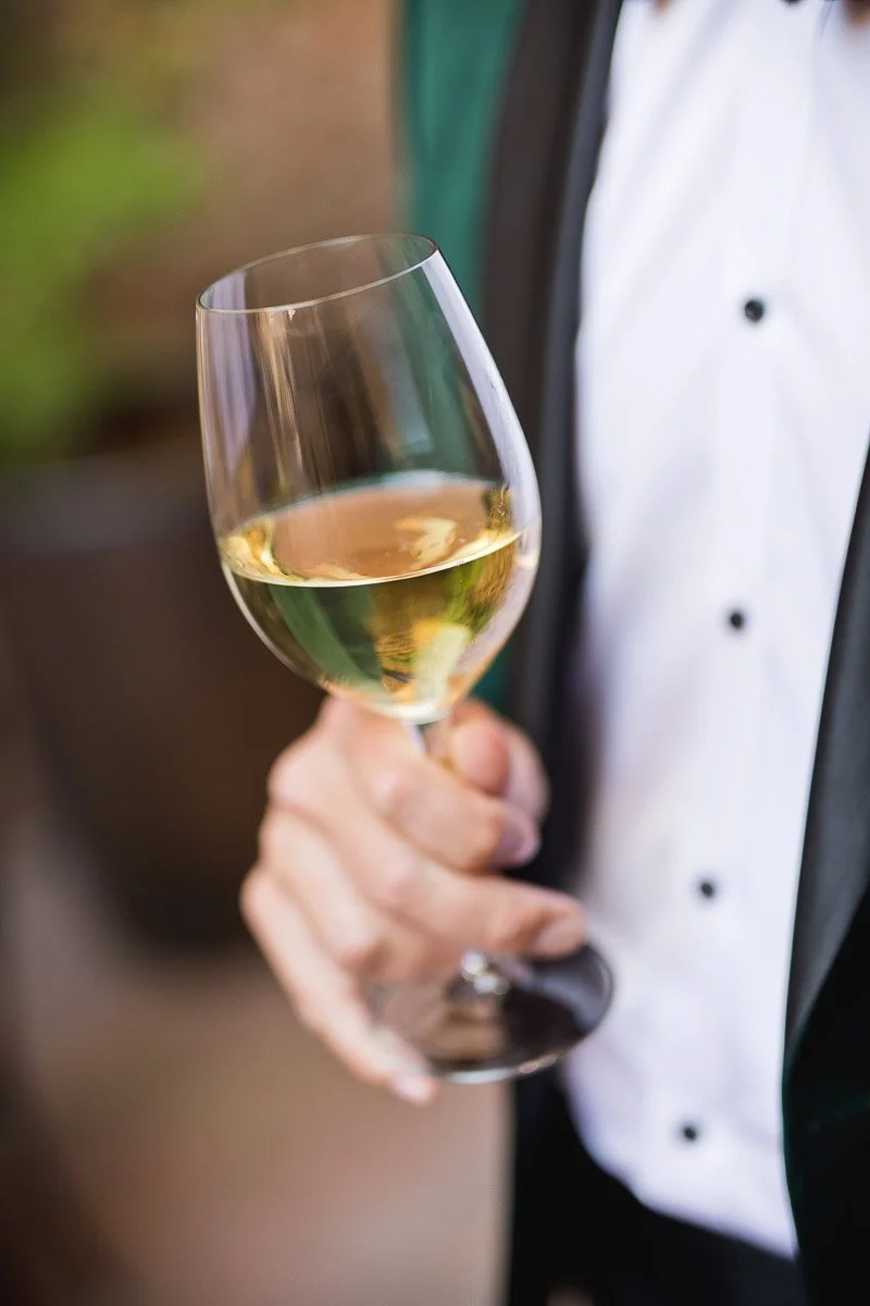 A hand holding a glass of white wine, with a person in formal attire visible. The setting conveys a sophisticated and elegant atmosphere.