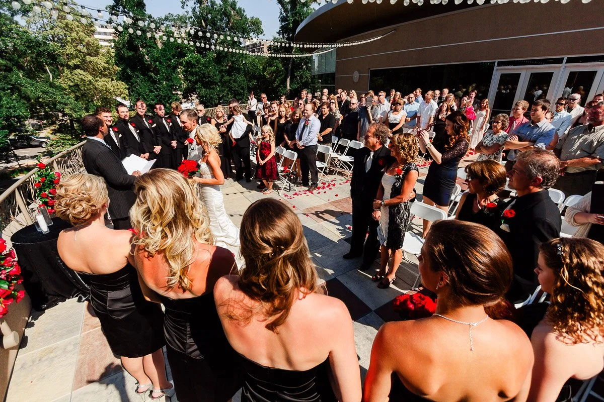 Elegant outdoor wedding ceremony with bride, groom, and officiant on a sunlit terrace. Guests in formal attire observe, surrounded by greenery and festive decor.