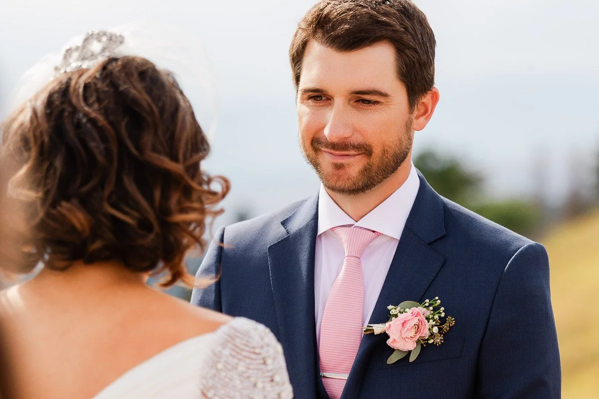 A groom in a blue suit with a pink tie and boutonniere gazes warmly at a bride, who is slightly turned away, in an outdoor, romantic ceremony setting.