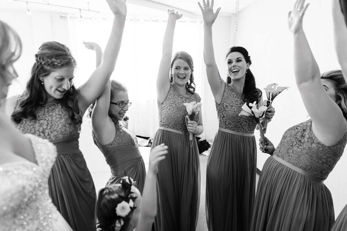 Bridesmaids in lace dresses raise their arms joyously, holding bouquets. They stand in a bright room, exuding excitement and celebration.