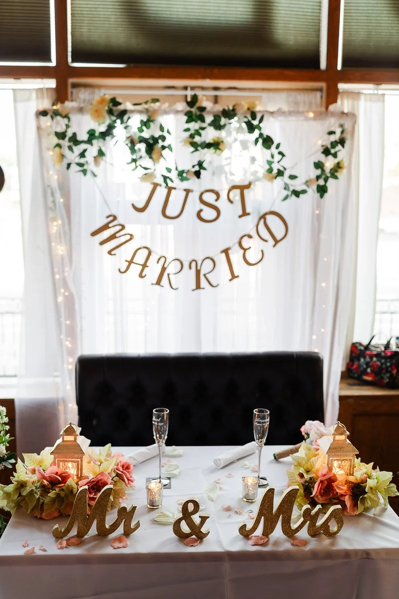 Wedding table setup with a "Just Married" banner, floral garlands, glowing lanterns, and "Mr & Mrs" signs, conveying a joyous, romantic ambiance.