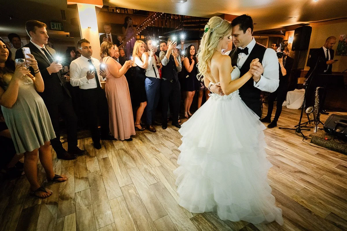 A bride and groom share their first dance in a warmly lit room, surrounded by smiling guests capturing the moment with phones, creating a joyful atmosphere during a The Lobby wedding reception in Denver