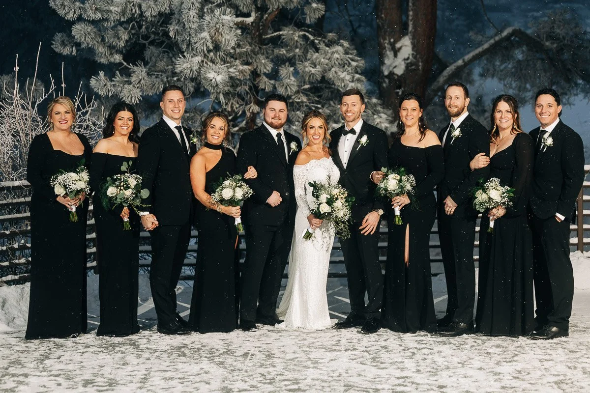 A wedding party stands in a snowy landscape. The bride in a white gown is surrounded by bridesmaids in black dresses and groomsmen in black suits, all holding bouquets. They radiate joy against a backdrop of snow-covered trees.