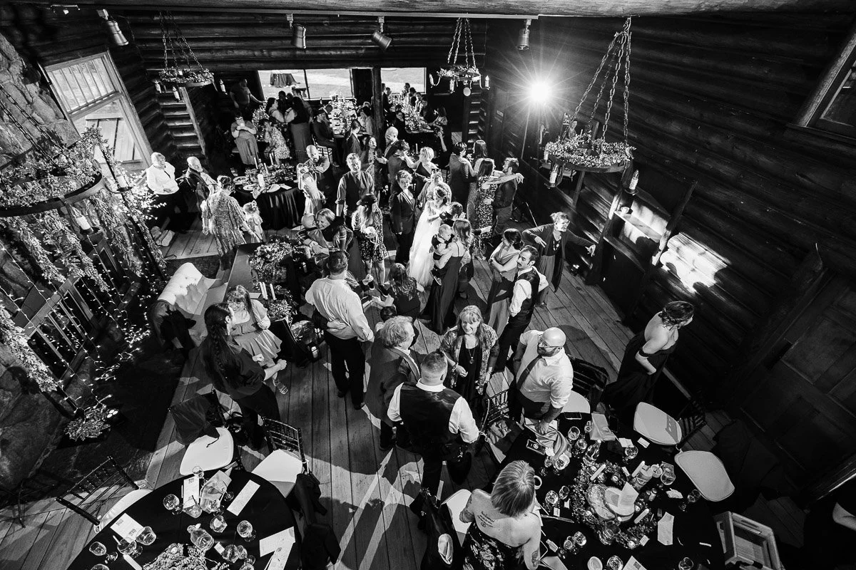 A black-and-white aerial view of a lively wedding reception in a rustic venue. Guests are dancing and mingling under soft lights, creating a joyful atmosphere.