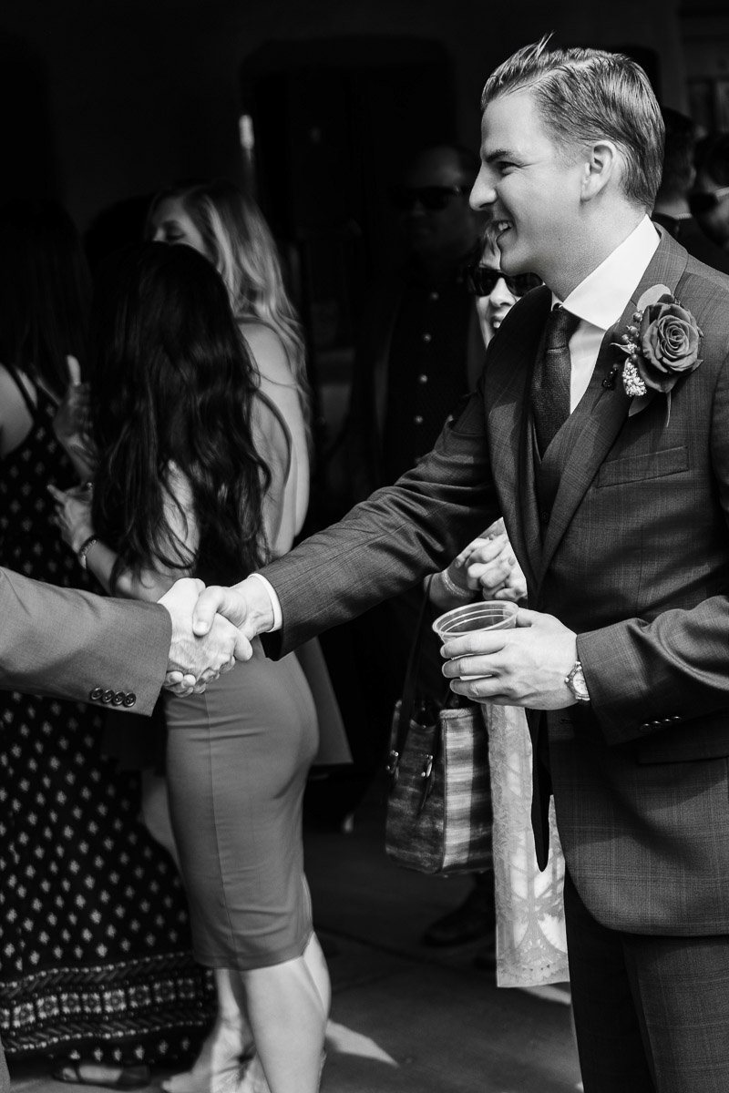 A man in a suit, with a flower boutonniere, shakes hands at a social event, holding a cup. Guests, some chatting and dressed in formal attire, fill the background.
