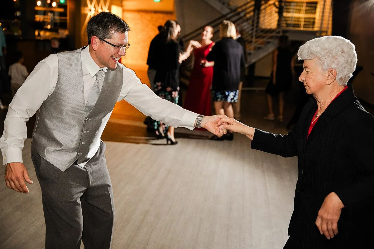 A man in a gray vest and tie dances joyfully with an elderly woman in a black suit. Background shows people socializing in a warmly lit room.