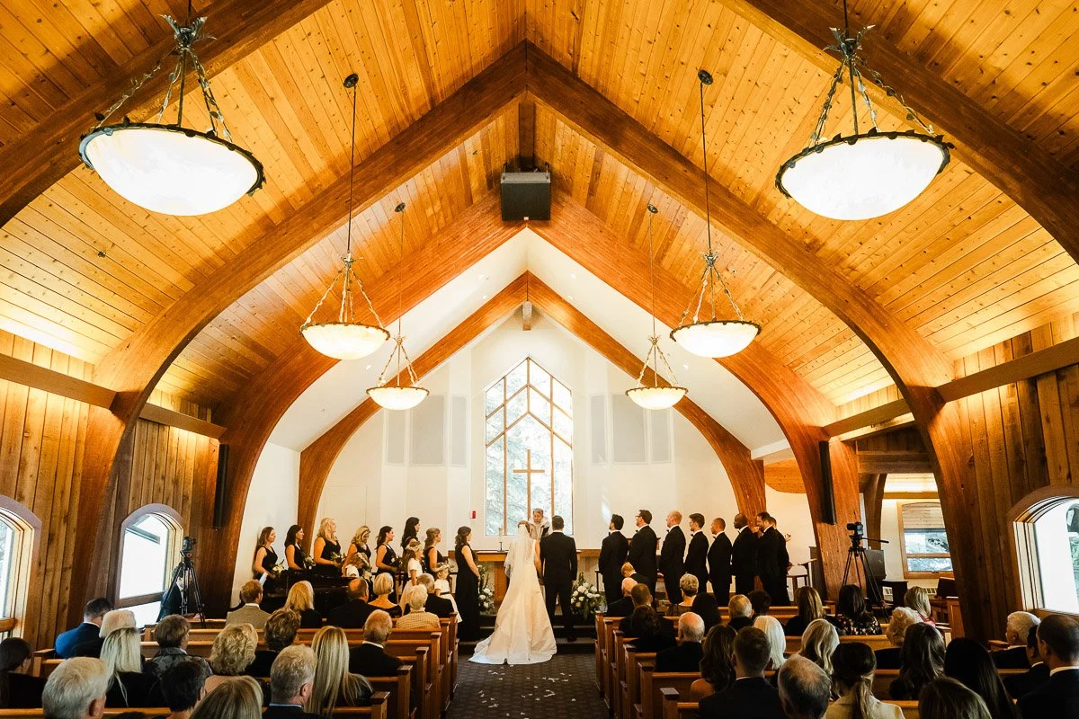 Bride and groom at altar in Vail Interfaith chapel, surrounded by bridesmaids and groomsmen. Guests seated, warm lighting, and serene ambiance.
