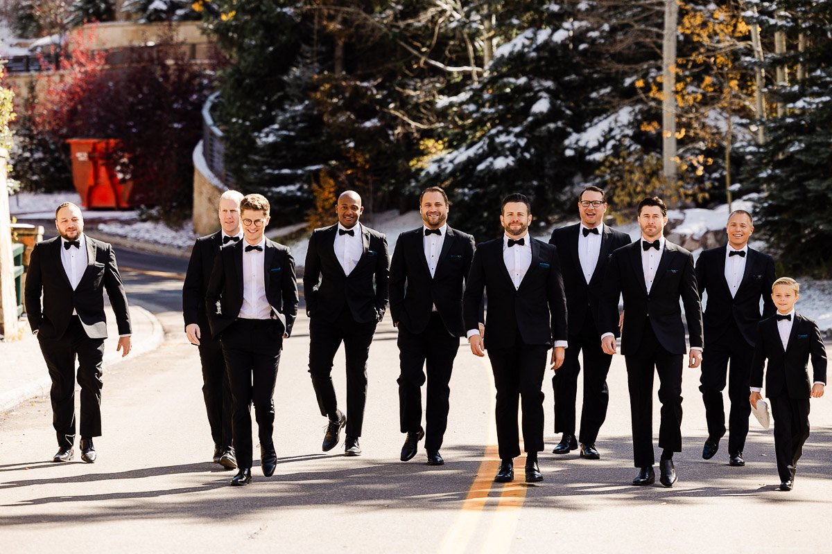A group of nine men and one boy in tuxedos walk confidently down a sunlit road. Snow-dusted trees and autumn leaves form a vibrant backdrop.