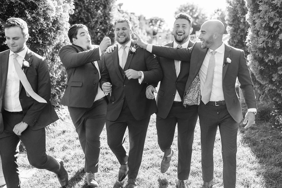 Five men in suits walk outdoors, laughing and joking, on a sunny day. They exude camaraderie and joy, surrounded by greenery. Black and white image.