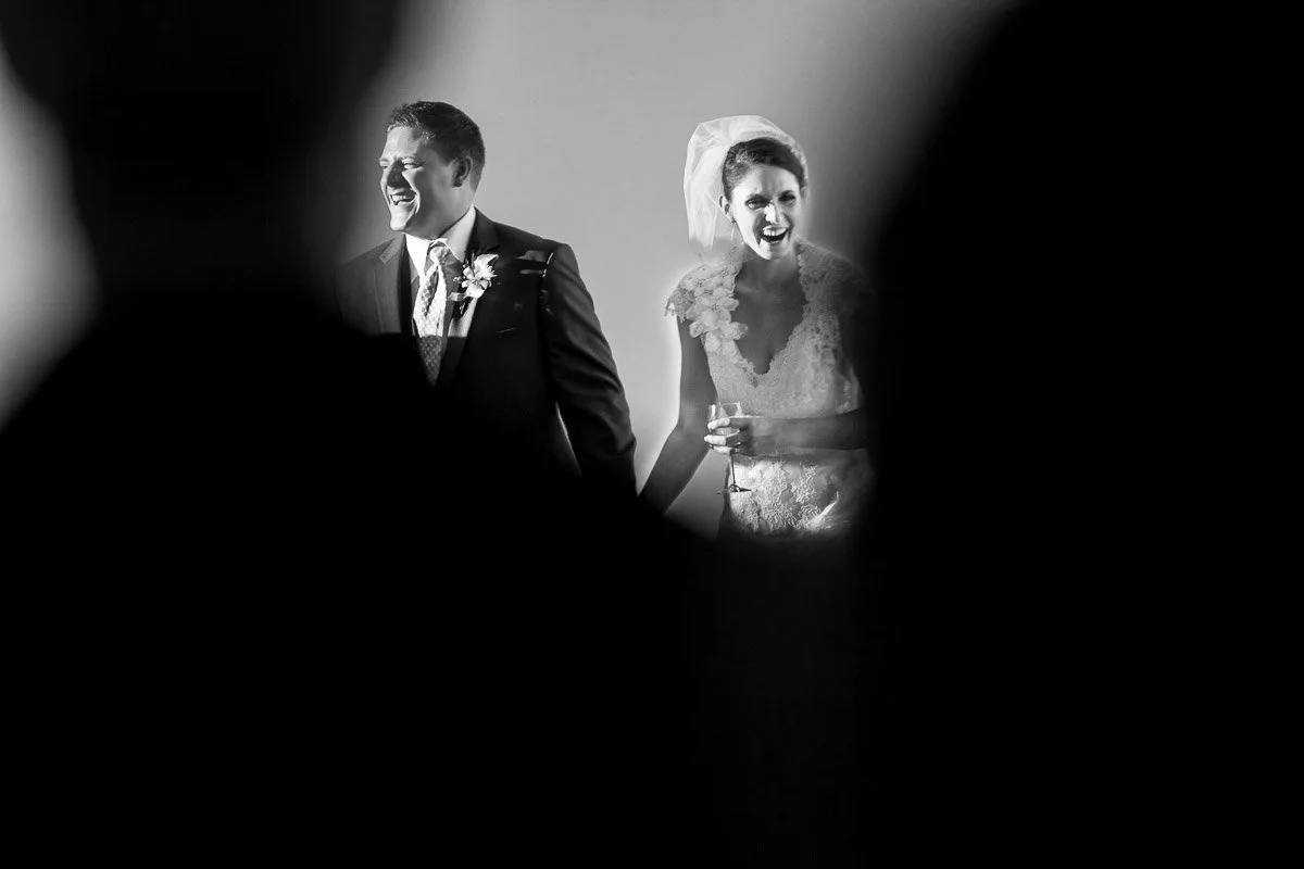 A joyful bride and groom are captured in a candid moment, holding hands and smiling, with soft lighting highlighting their expressions. The grayscale tones add a classic, romantic feel.