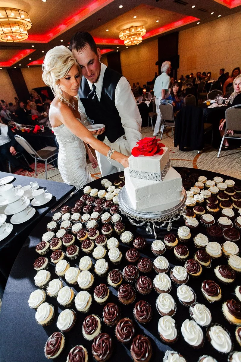 Bride and groom cutting a square white wedding cake with a red rose on top, surrounded by black and white cupcakes. Elegant setting, warm ambiance.