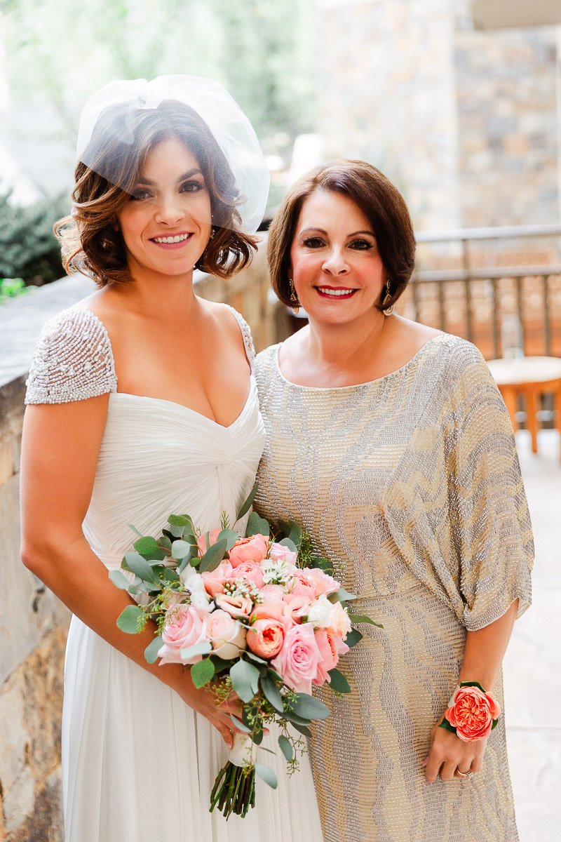 A bride in a white gown with embellished sleeves holds a bouquet of pink roses. She stands beside a smiling woman in a silver dress. Both appear joyful.