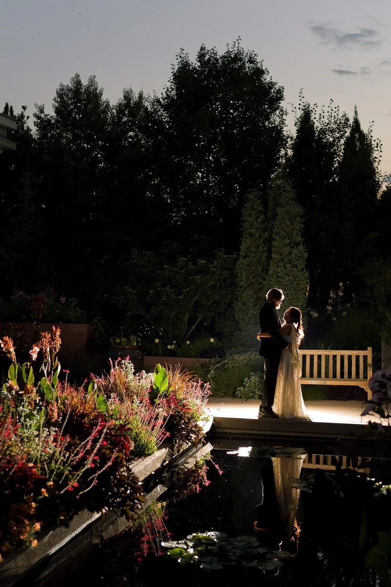 A couple embraces on a garden path at dusk, illuminated by soft lighting. Lush plants and a serene pond add romance to the tranquil scene.
