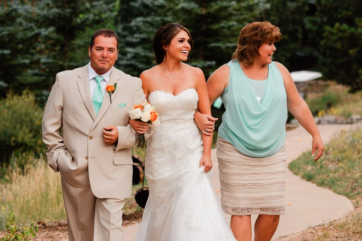 A smiling bride in a white lace gown walks arm-in-arm between a man in a beige suit and a woman in a teal top, all on a garden path.