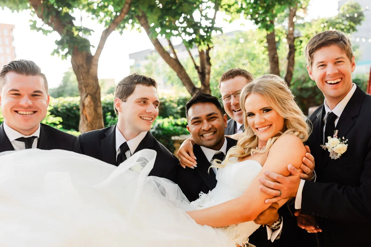 A joyful bride in a white gown is lifted by five smiling groomsmen in black suits and ties. They stand outdoors, surrounded by lush green trees.