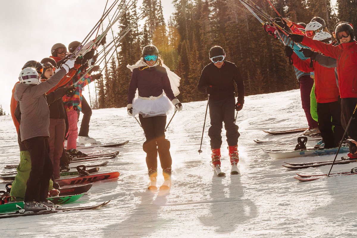 A skier in a tutu glides down a snowy slope, flanked by cheering friends holding ski poles aloft. Sunlight filters through trees, creating a joyful scene.