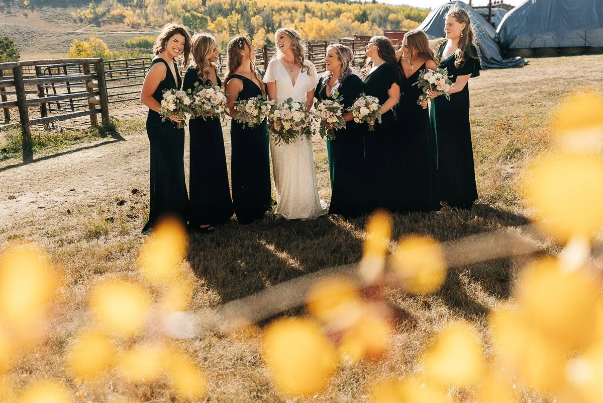 A bride in white and seven bridesmaids in dark dresses stand joyfully together outdoors, holding bouquets. Sunlight and autumn trees create a warm atmosphere.