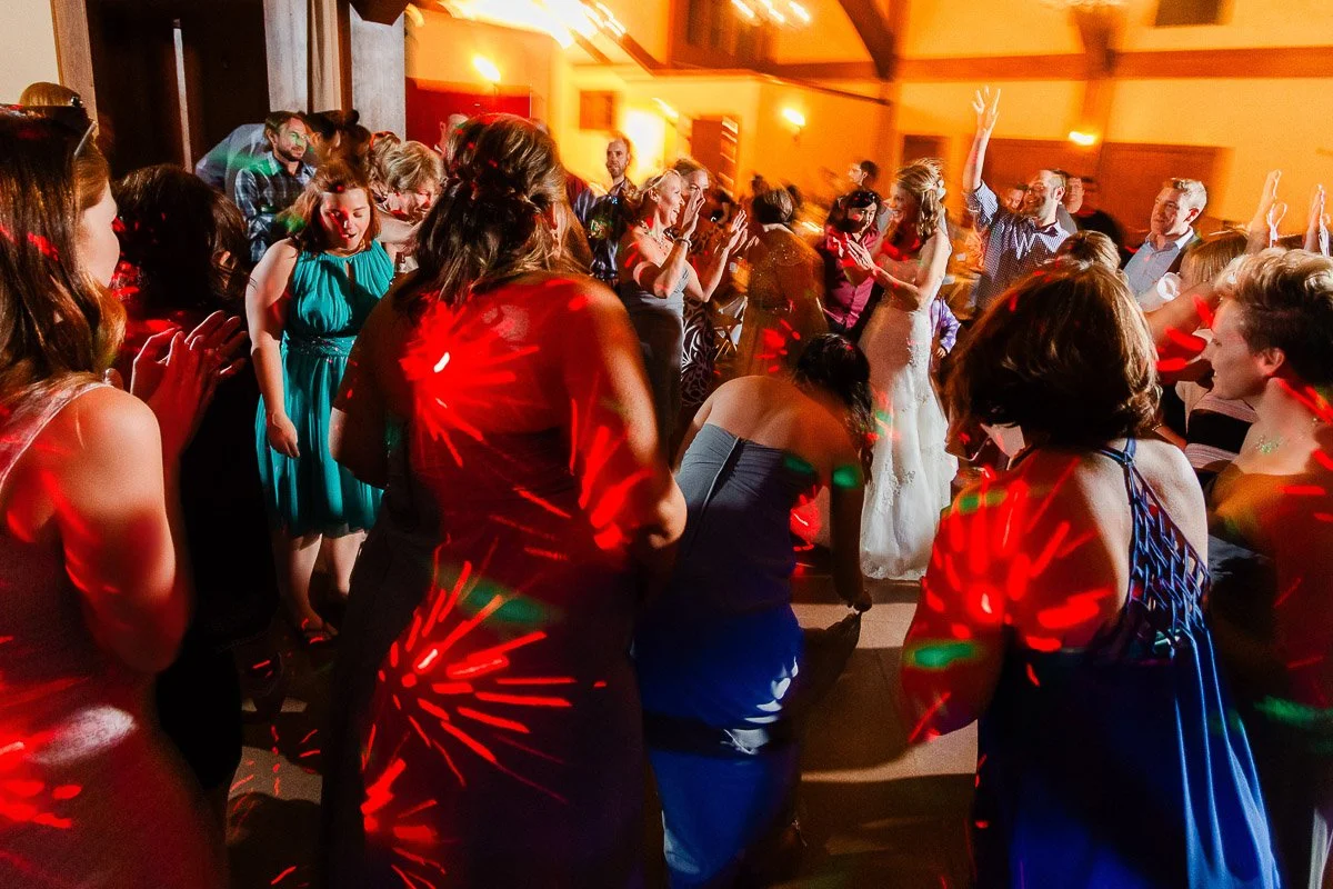 A lively dance floor scene at a wedding with colorful lights casting patterns. Guests in formal attire are dancing energetically and clapping.
