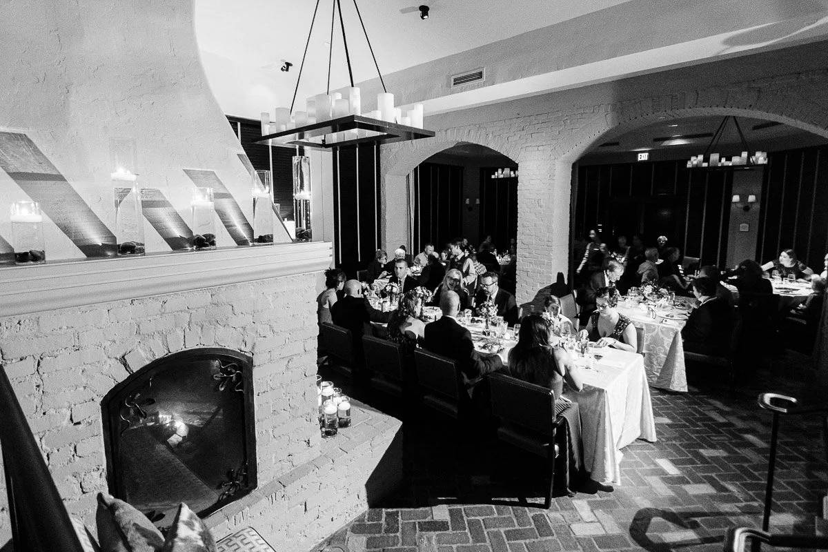 Black and white photo of an elegant candle-lit dinner in a rustic room with a fireplace. Guests in formal attire are seated, creating a warm, intimate atmosphere.