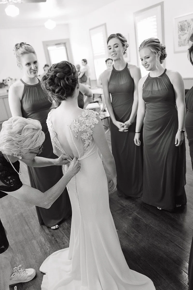 A bride in an elegant gown has her dress adjusted by an older woman while three bridesmaids in matching dresses watch with warm smiles. The scene is light-hearted and joyful.