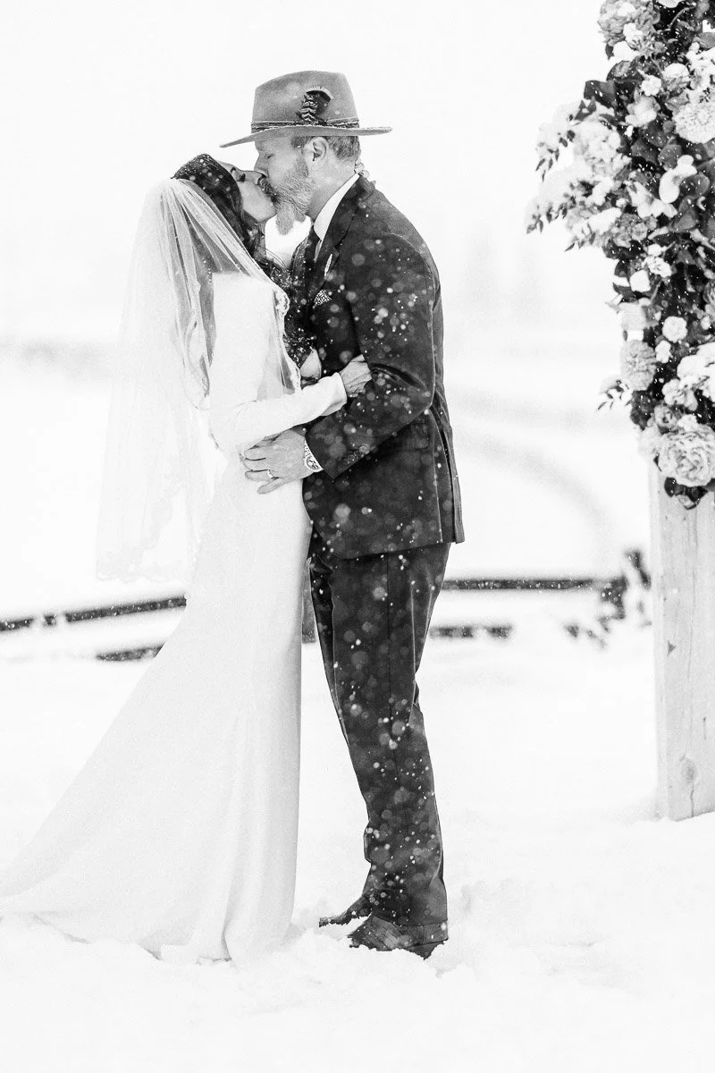 Bride and groom kissing in a snowy outdoor setting, both in formal attire, under a floral arch. Snowflakes gently fall around them, creating a romantic atmosphere.