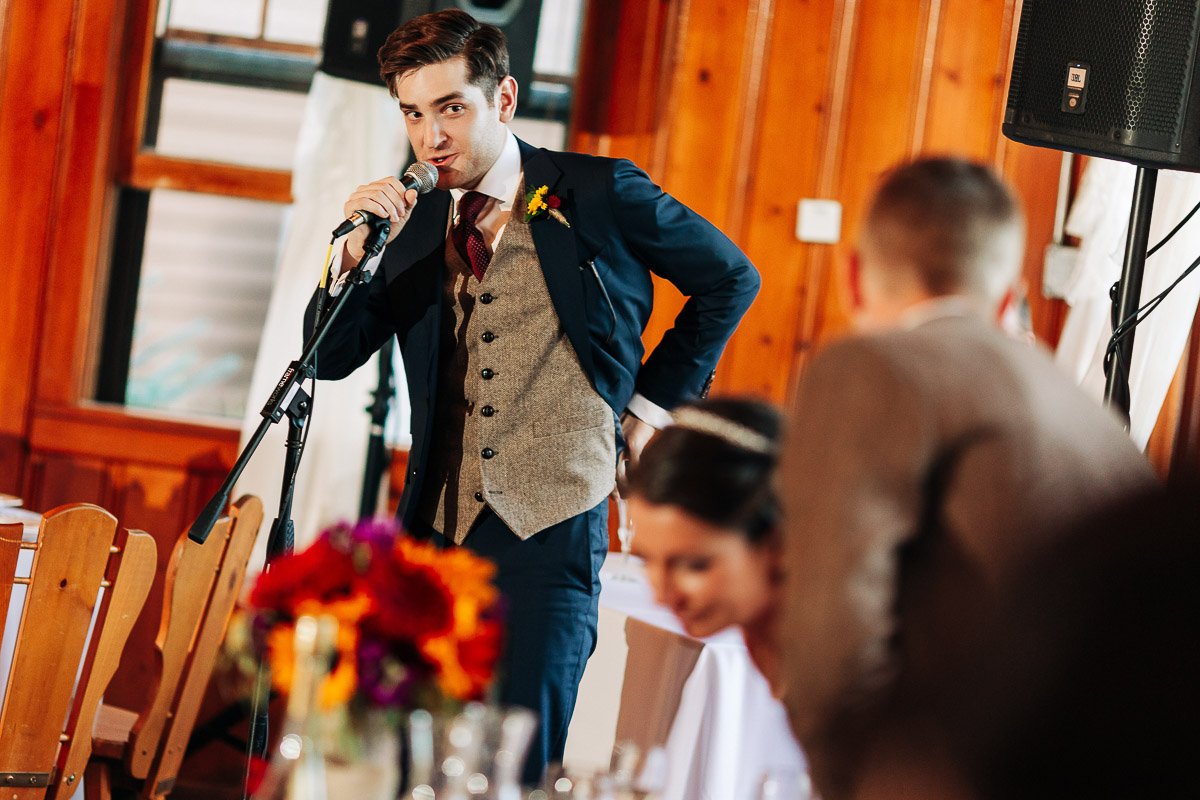 A man in a suit with a flower boutonniere speaks into a microphone at an indoor event. People in the foreground, a vibrant bouquet, and wood-paneled walls create a warm, joyful atmosphere.