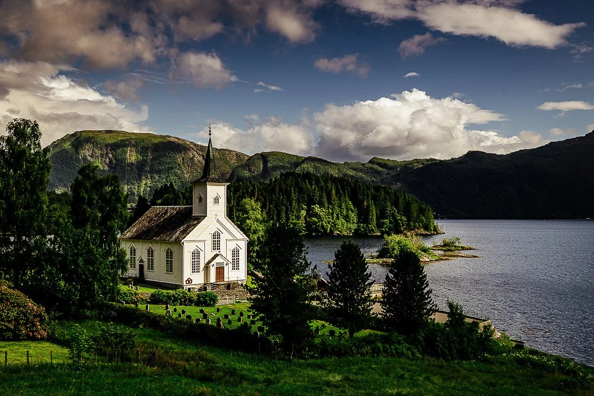 White church with a tall steeple set in lush greenery by a tranquil lake. Mountains and clouds in the background under a partly cloudy sky. Serene atmosphere.