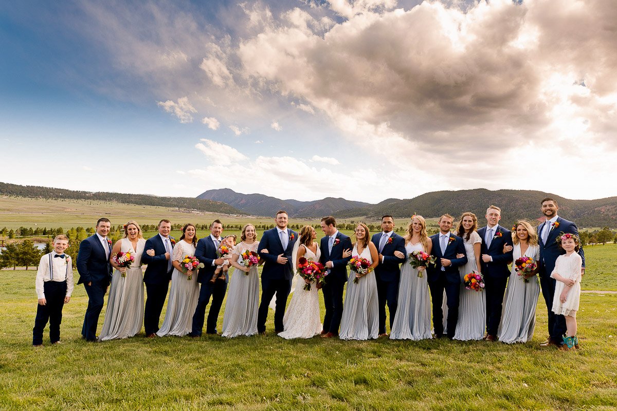 A smiling wedding party poses in a scenic meadow. Bridesmaids in gray dresses and groomsmen in blue suits stand under a cloud-filled sky. The mood is joyful during a Spruce Mountain Ranch wedding in Larkspur, Colorado