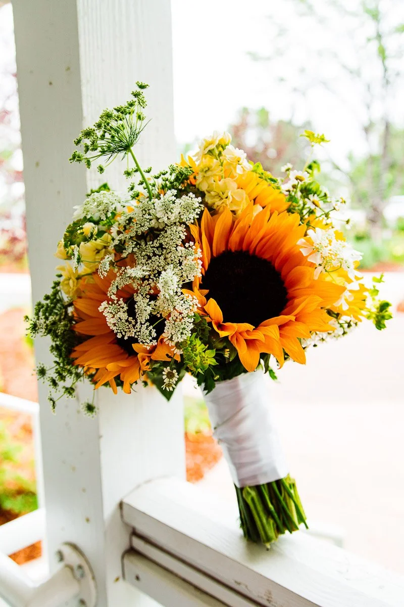 Bouquet with vibrant sunflowers, delicate white flowers, and greenery, wrapped in white ribbon, resting on a white porch railing, conveying a cheerful, rustic vibe.