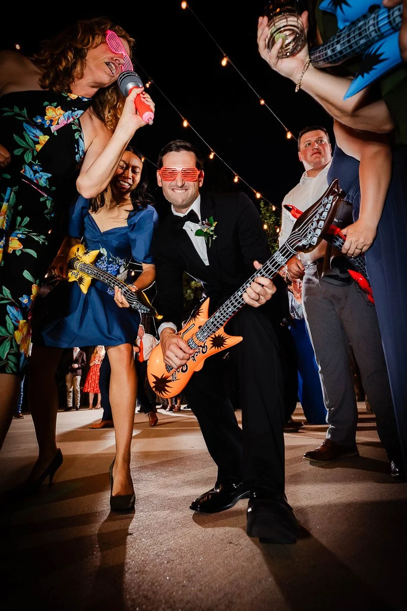 A joyful group dances with inflatable guitars under string lights. They wear colorful sunglasses and party attire, creating a lively, festive atmosphere.