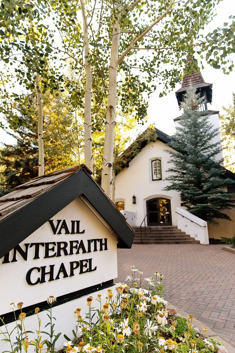 White chapel with a steep roof and bell tower, surrounded by tall trees in sunlight. Foreground sign reads "Vail Interfaith Chapel," with flowers below. Peaceful ambiance.