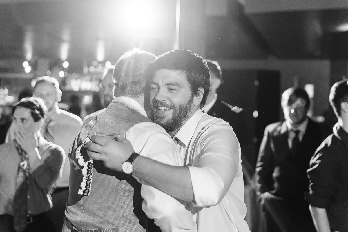 A joyous moment as two men embrace in a warmly lit event, surrounded by smiling guests. The black-and-white photo exudes happiness and camaraderie.