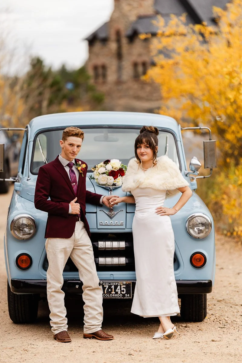 A wedding couple poses in front of a vintage light blue Dodge truck. The groom wears a burgundy blazer; the bride wears a white dress, conveying a retro, cheerful vibe captured by Estes Park wedding photographer tomKphoto