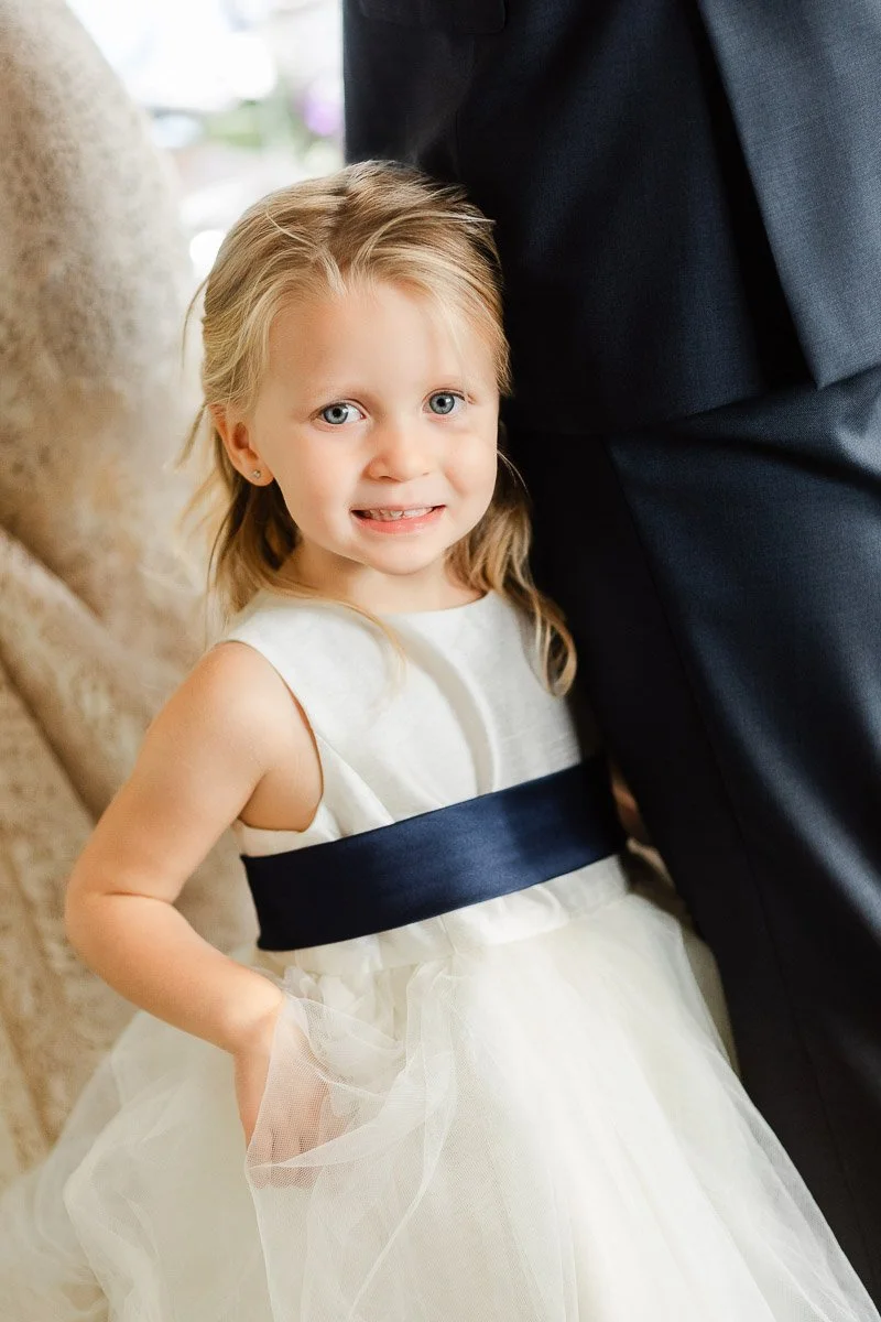 A young girl in a white dress with a navy sash, smiling and standing beside an adult in a dark suit. The scene conveys warmth and joy.