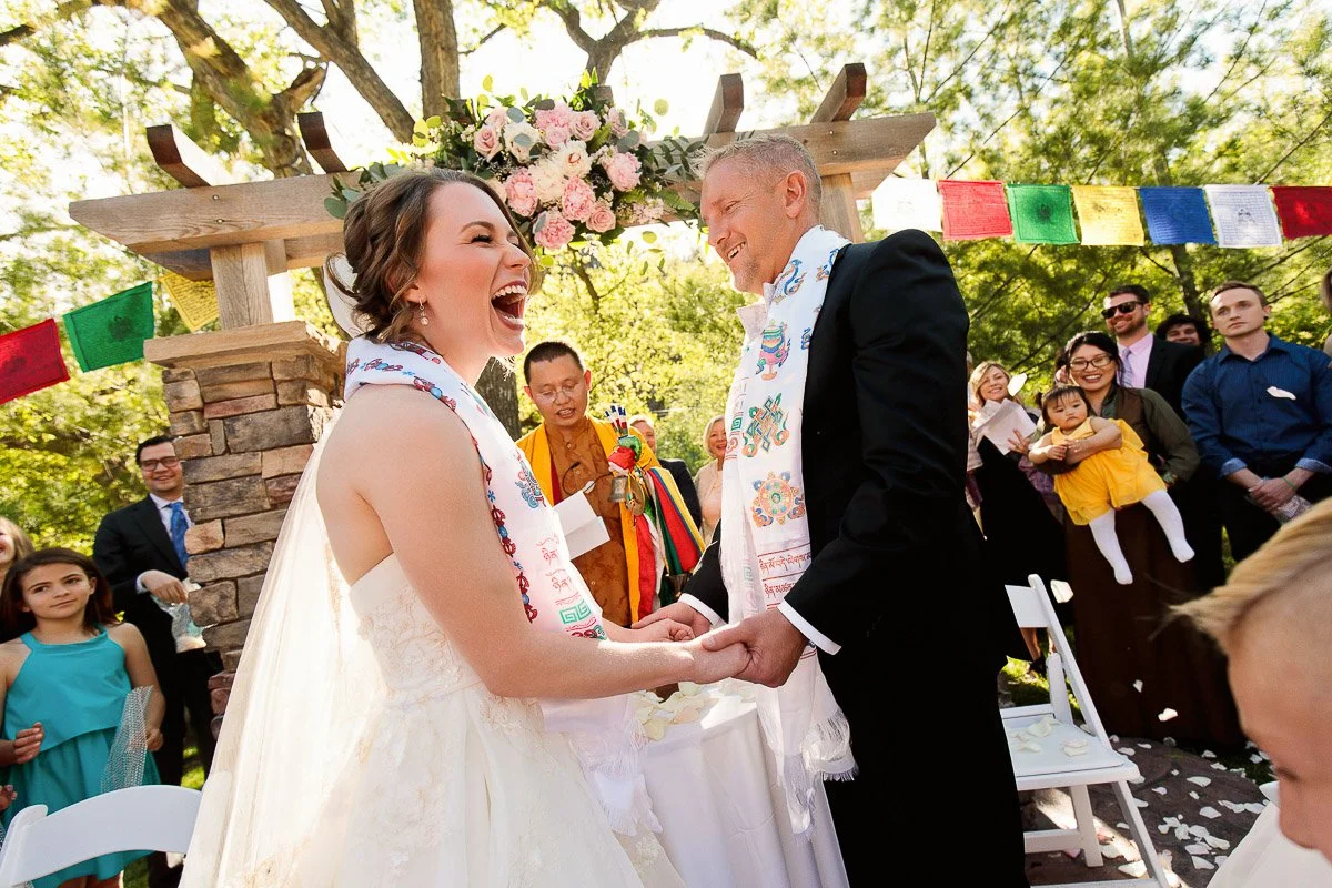 A joyful wedding scene shows a bride and groom laughing and holding hands under a floral arch. Guests, including a Buddhist monk, look on happily.