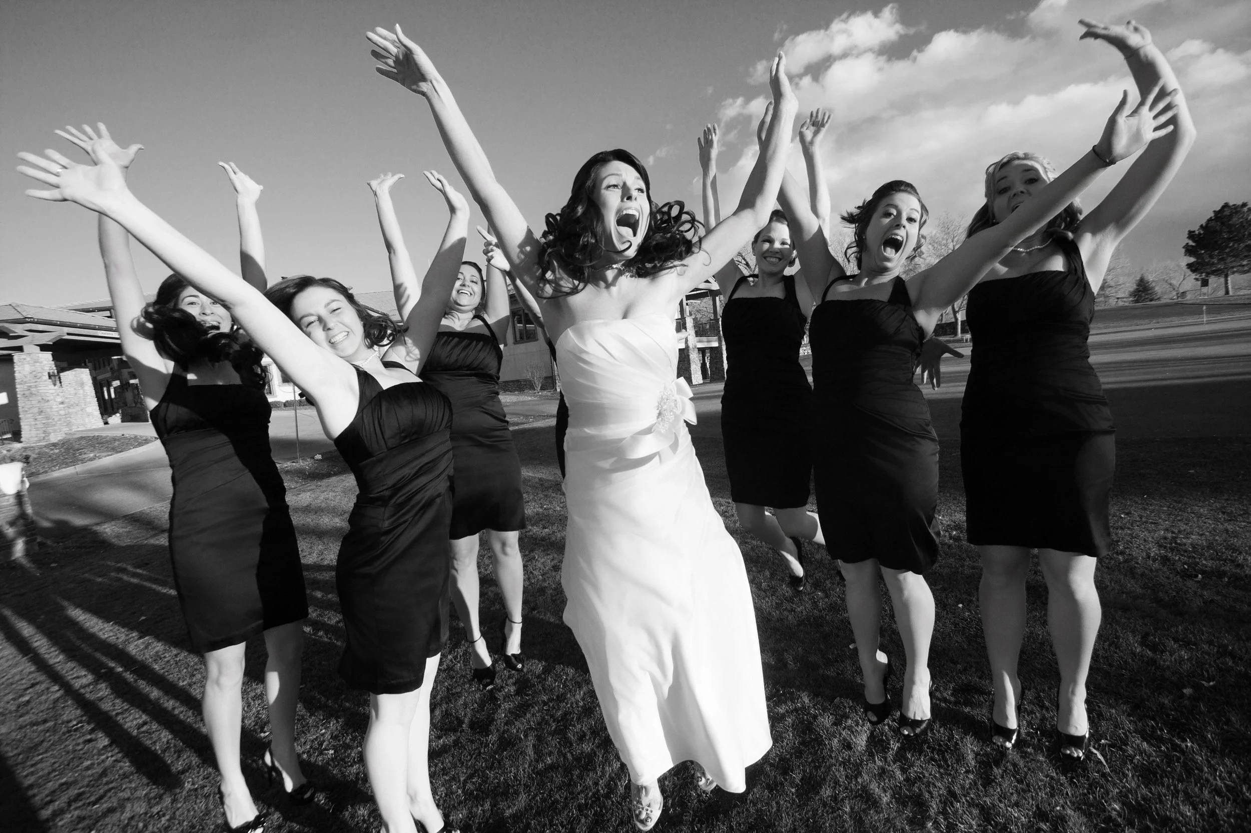 A joyful bride in a white dress surrounded by seven bridesmaids in black dresses, all with arms raised, laughing and jumping against a bright sky.