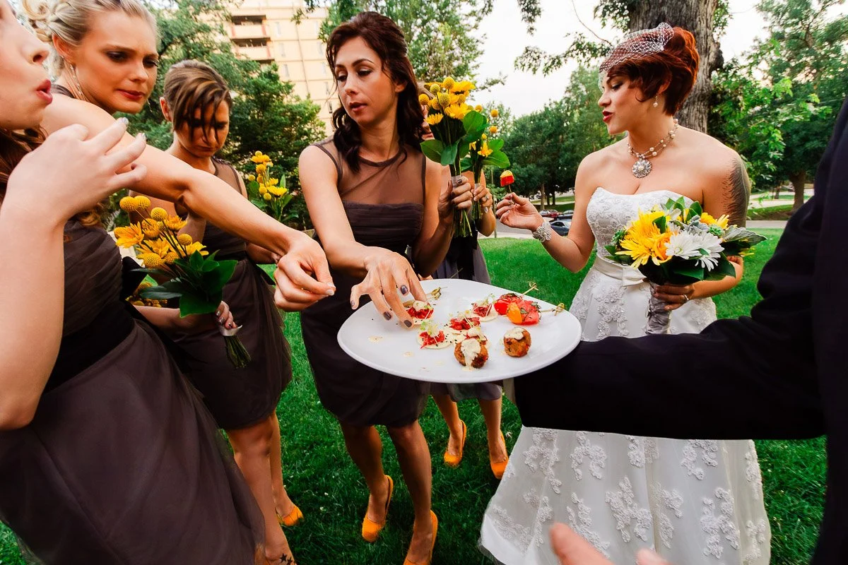 Bridesmaids in dark dresses eagerly reach for appetizers from a plate held by a person in black; the bride in white holds a bouquet, smiling.