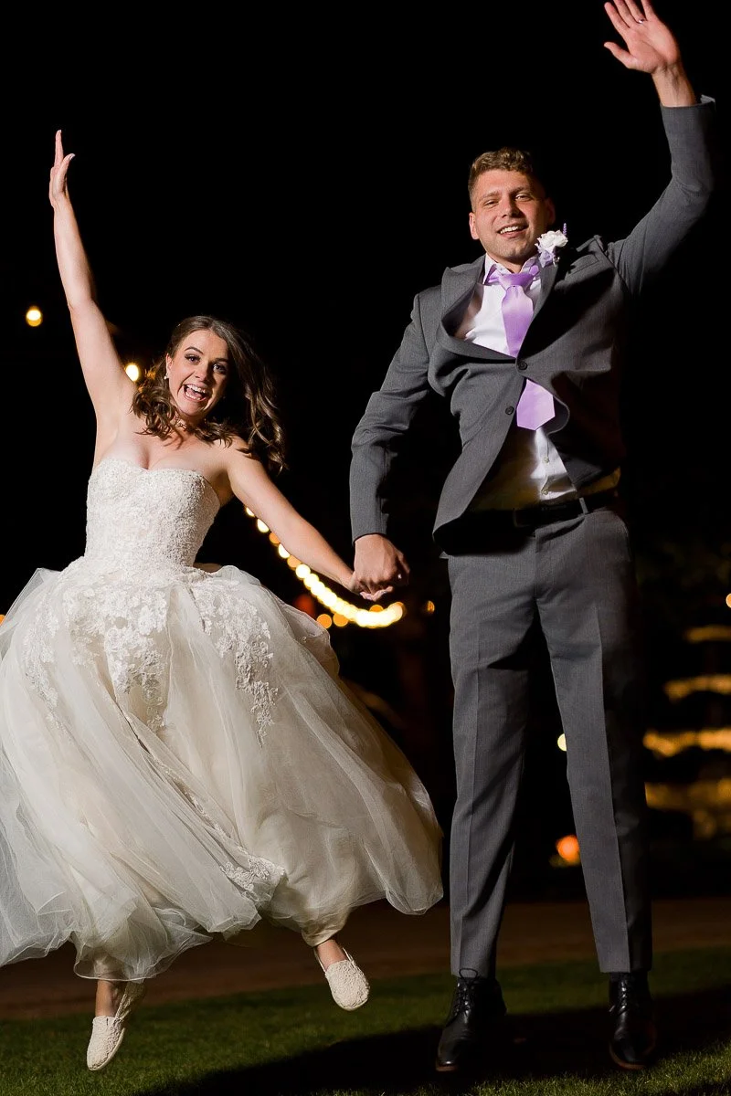 Bride and groom joyfully jump in the air at night, holding hands. The bride wears a flowing white gown; the groom, a gray suit with a lilac tie. Festive lights glow in the background during a Church Ranch Event Center wedding in Westminster, Colorado