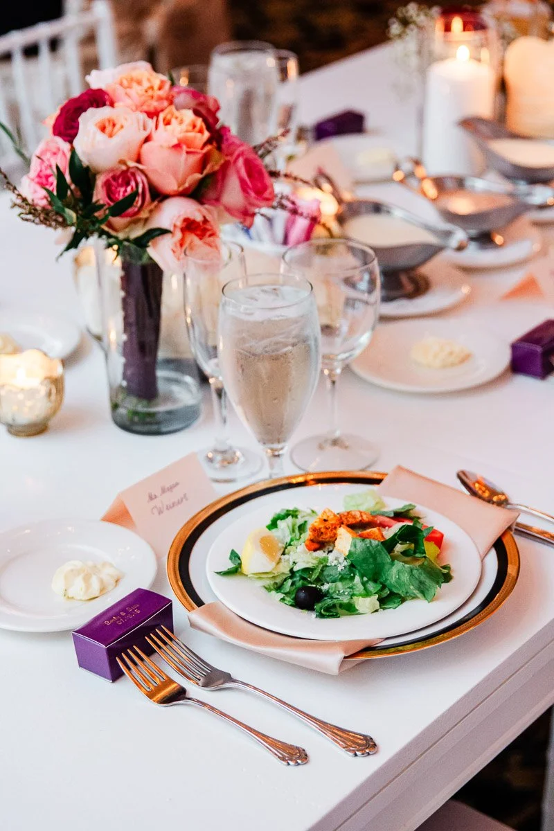 Elegant table setting with a fresh salad on a white plate, pink rose centerpiece, and a glass of water. Warm, celebratory atmosphere.