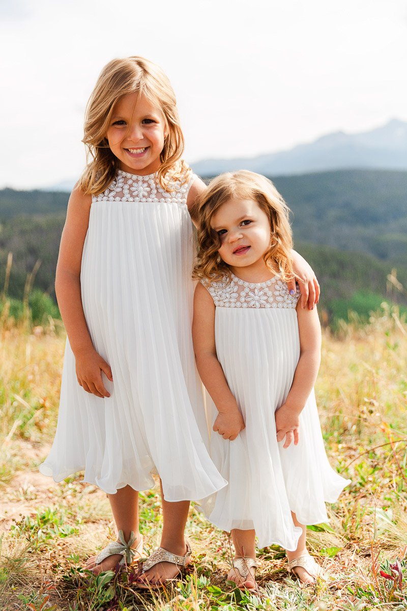 Two young girls in white dresses stand joyfully in a sunny meadow, with one arm around each other. The background shows green hills and a distant mountain.