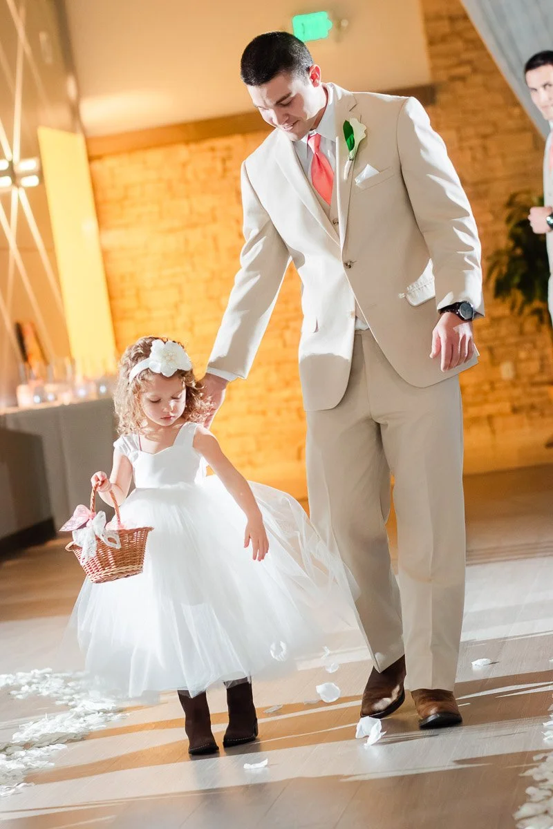 Man in beige suit walks with a young girl in a white dress holding a basket, scattering petals. Warm lighting creates a joyful, celebratory scene.