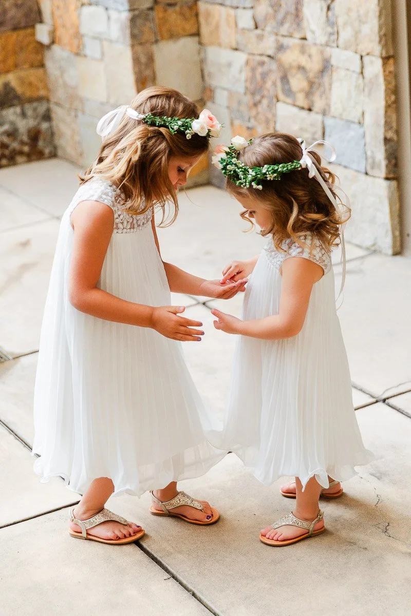 Two young girls in white dresses and floral headbands stand on a patio, gently holding hands. The scene is lighthearted and tender.