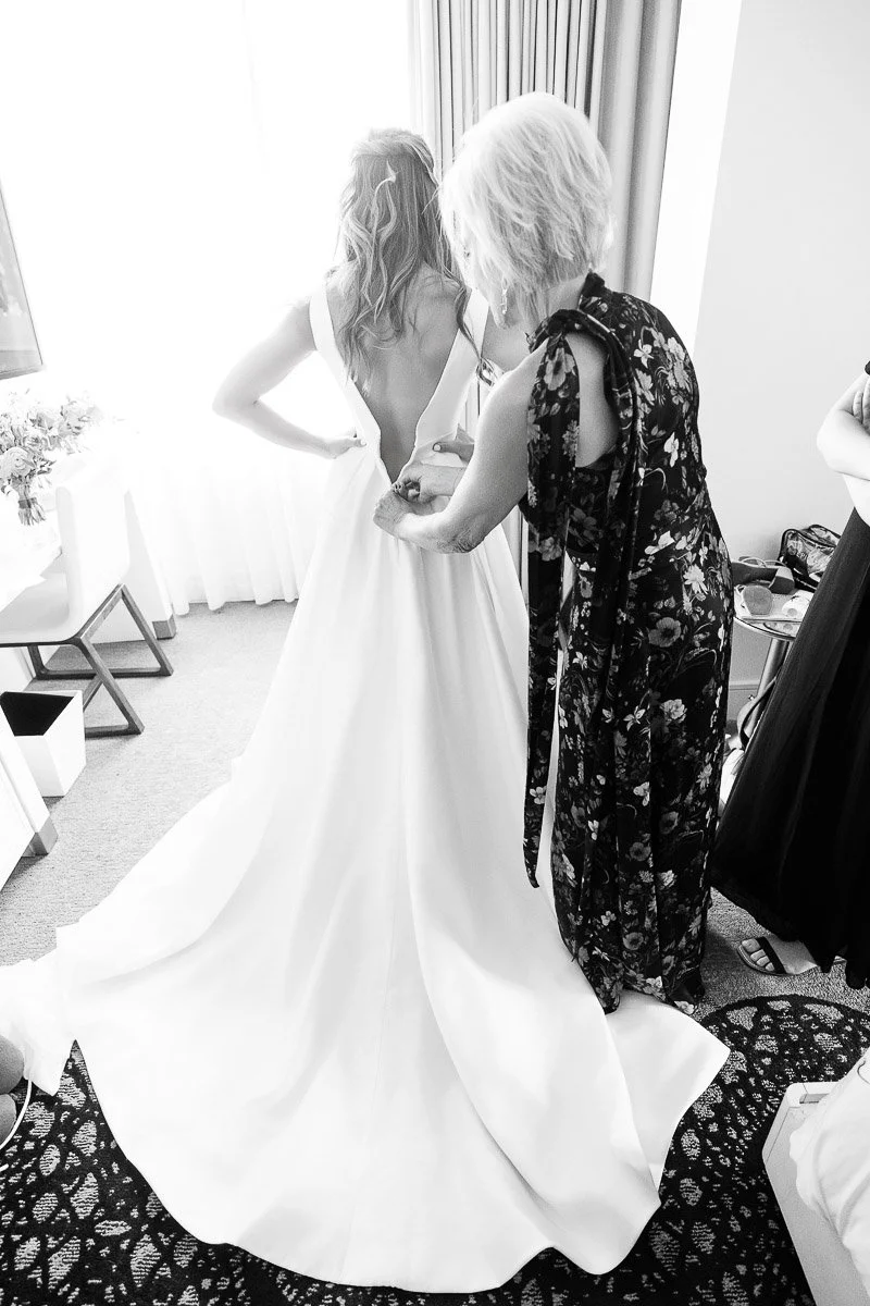 A bride in a flowing white gown stands as another woman assists with adjustments. The mood is intimate, with soft light from a nearby window.