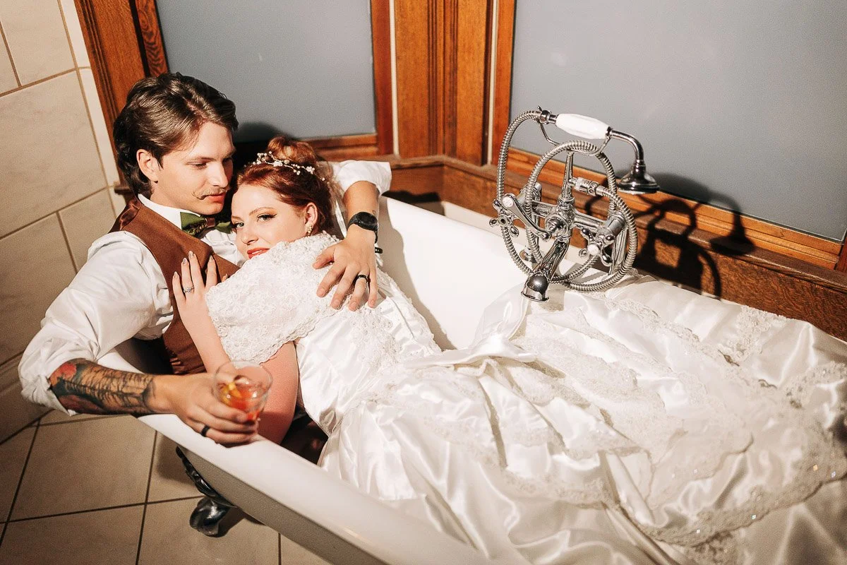 A bride and groom in wedding attire are lounging playfully in a white clawfoot bathtub. The groom holds a drink, and they both look relaxed and happy.