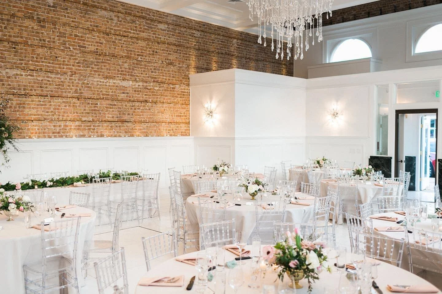 Wedding reception setup at The Treasury wedding venue in Delavan Wisconsin featuring exposed brick walls, crystal chandeliers, and clear chiavari chairs.