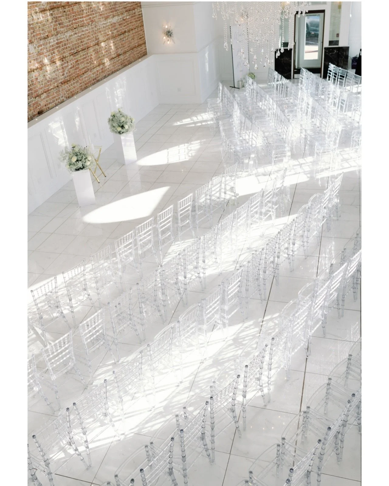 Indoor wedding ceremony setup at The Treasury wedding venue in Delavan Wisconsin featuring crystal chandeliers, white floors, exposed brick wall, and clear chiavari chairs.