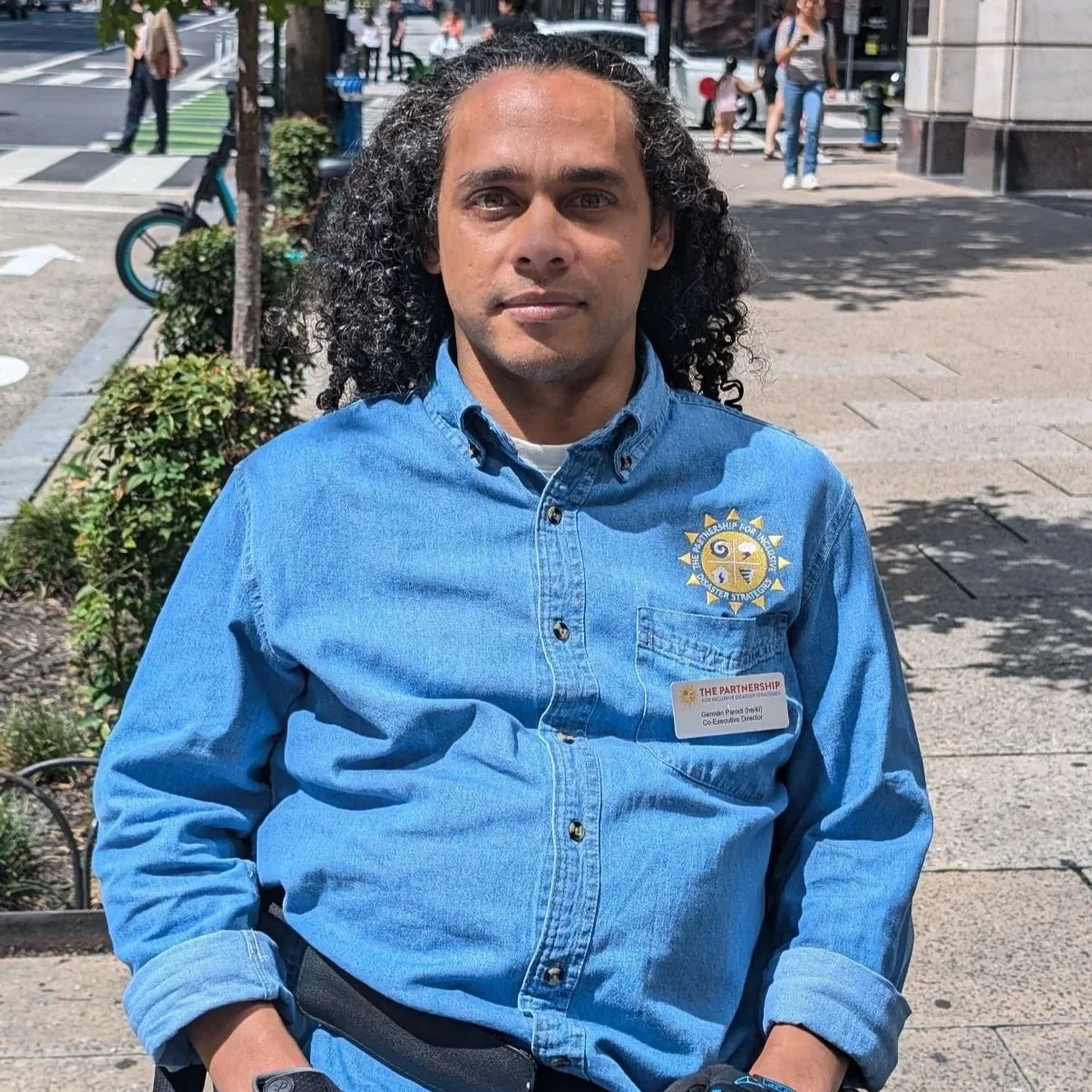 Image of Germán, a Puerto Rican man with long curly hair sitting in his wheelchair on a city sidewalk, wearing a denim button-down shirt with The Partnership for Inclusive Disaster Strategies logo on it.