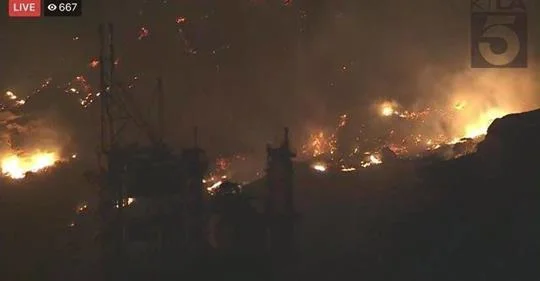 [Image Description: This photo has a dark background with two clusters of fire and smoke throughout the image. In the lower left-hand side of the image is a piece of infrastructure. In the upper right-hand corner is a “5” outlined by a black backgro…