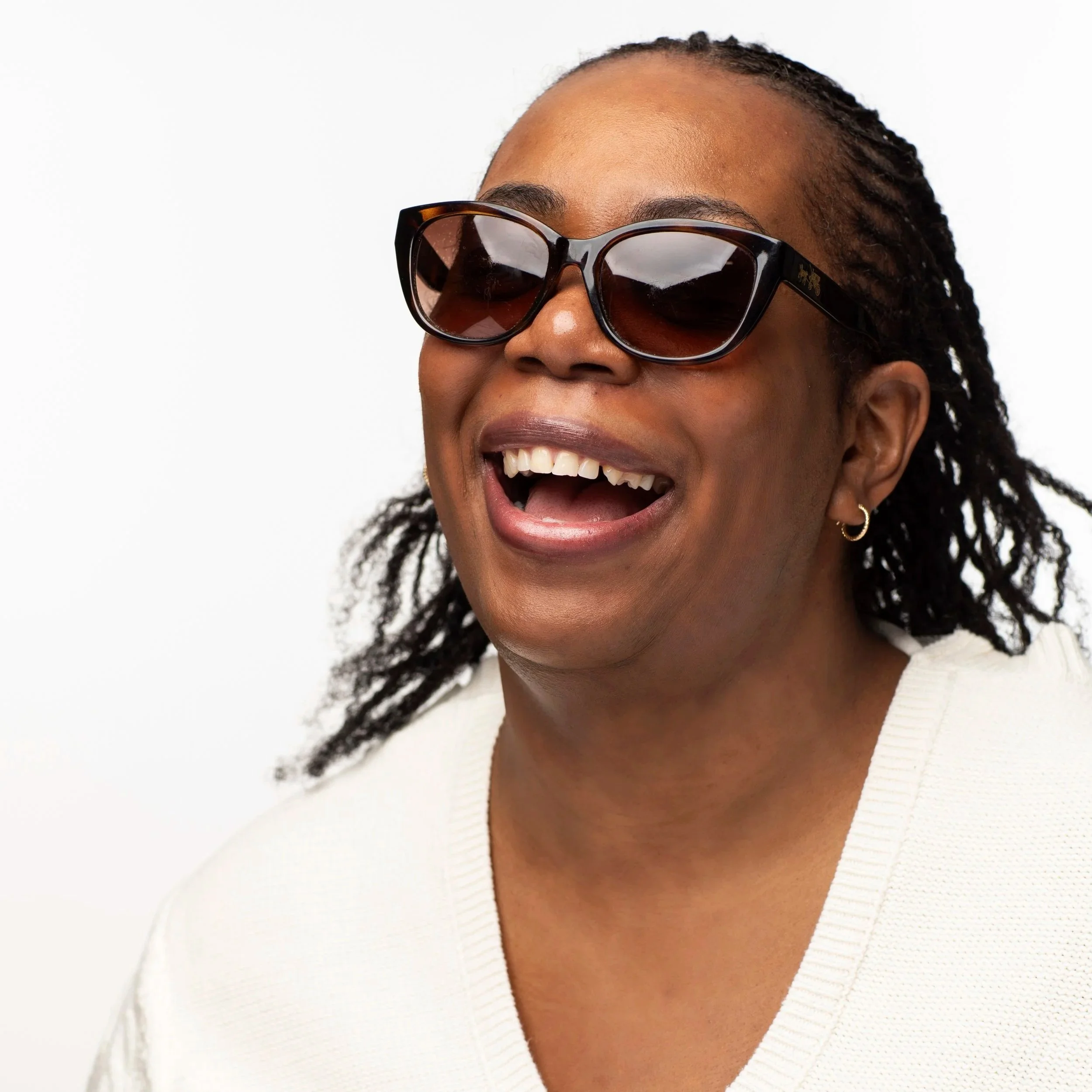 Justice, a Black blind woman with her hair styled in 2-strand twists, smiles joyfully for the camera wearing black sunglasses and a cream hooded sweater.