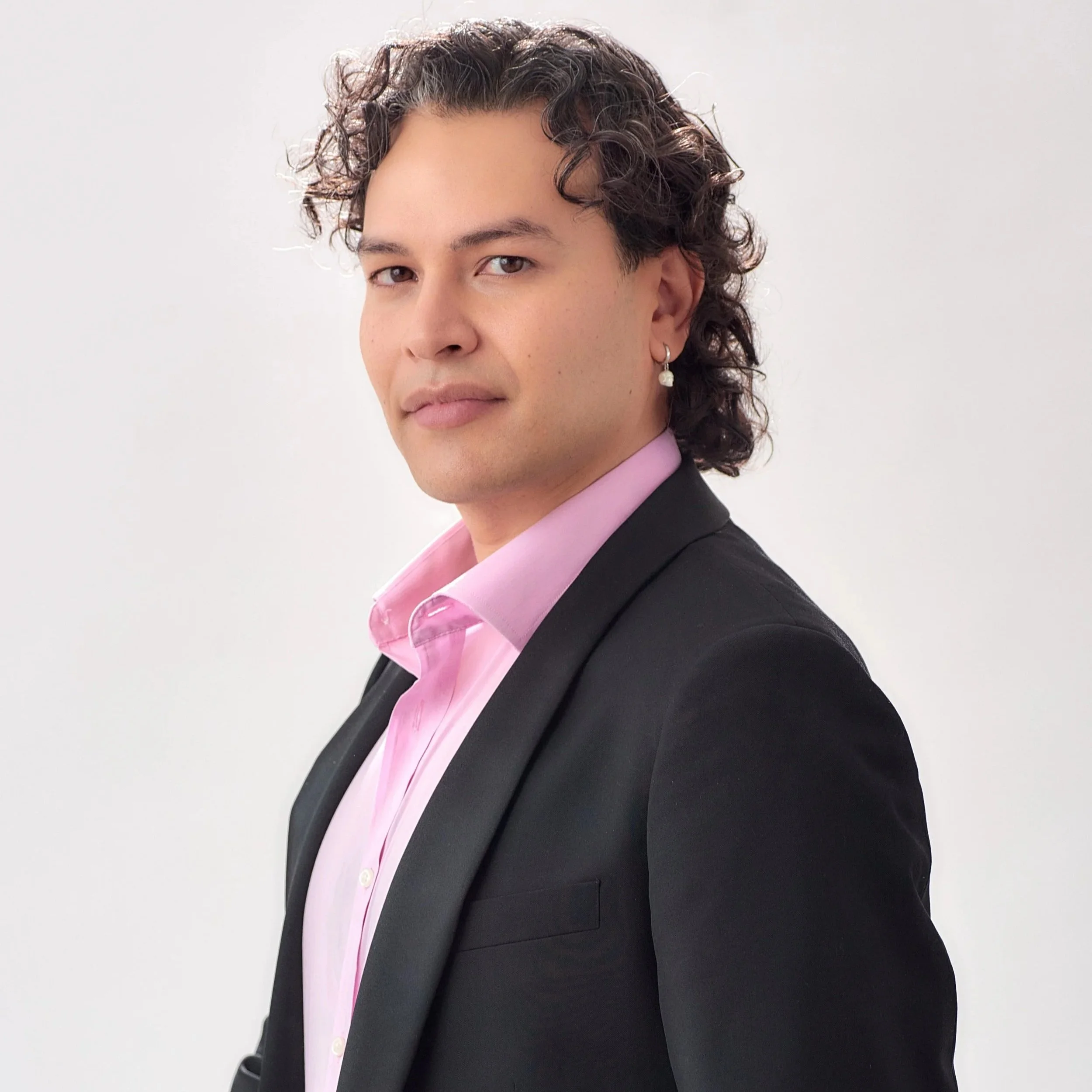 Image of Gabriel, a Mexican person with short curly dark hair sitting against a white background, wearing a pearl earring, light pink button-down shirt, and a black jacket.